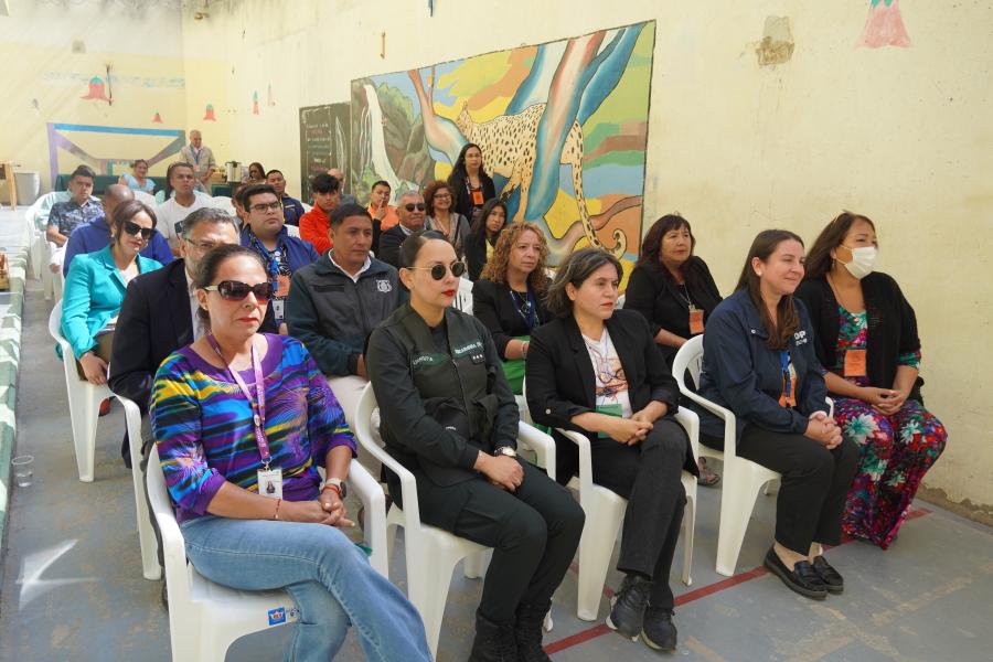 Un grupo de personas sentados en sillas de plástico blancas en un aula, escuchando atentamente a un orador fuera de cuadro, contra el telón de fondo de un mural colorido.