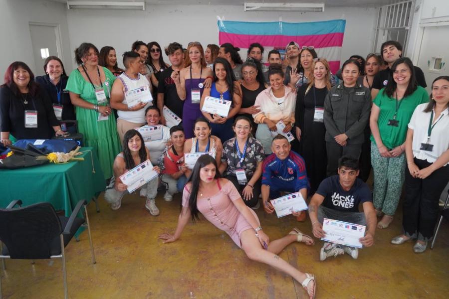 La imagen muestra un gran grupo de personas sentadas y de pie en una habitación sosteniendo su certificado de aprobación del curso. Al fondo hay una bandera de colores transgénero. 