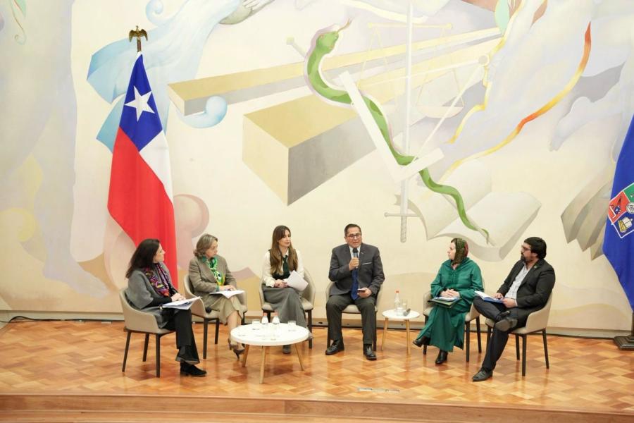 los cuatro panelistasy los dos moderadores sentandos en el escenario del salón de honor durante el panel, las sillas están dispuestas en forma de medialuna y atrás salen las banderas e Chile y de la Universidad de Chile